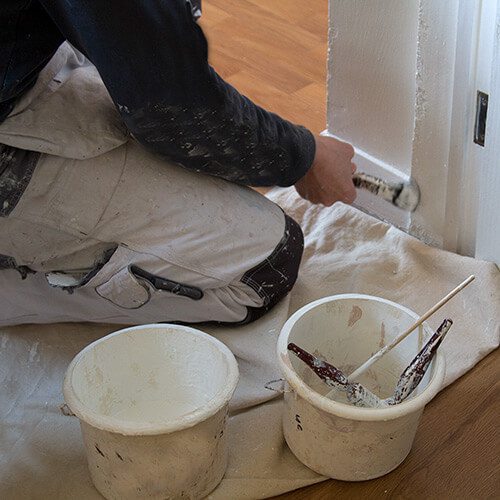 Painter kneeling on a drop cloth while painting white baseboard near a door frame with paintbrushes and paint containers nearby.