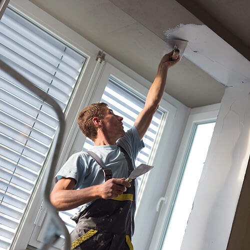 Man in work overalls applying plaster to a ceiling near windows with shutters.