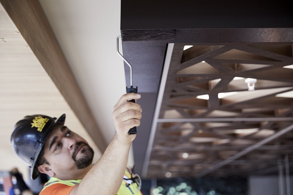 Construction worker wearing a hard hat and orange safety vest painting a metal ceiling frame with a paint roller.