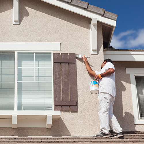 Painter in white clothing painting the exterior wall of a beige house near a window during daytime.