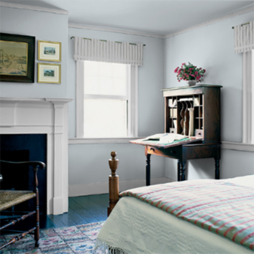 Cozy bedroom with a neatly made bed, vintage wooden desk with flowers, white fireplace, and framed artwork on light gray walls.