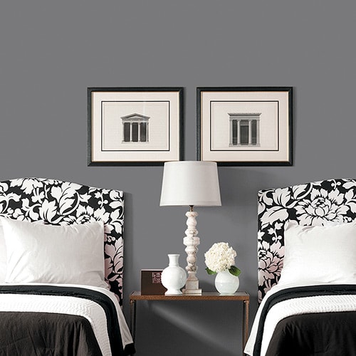 Two twin beds with black and white floral headboards separated by a wooden nightstand with a white lamp and decorative vases, against a gray wall with two framed architectural prints.