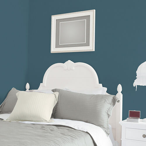 White vintage-style bed with gray and beige pillows against a teal wall with a framed geometric art piece above.