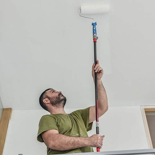 Man using a paint roller with an extension pole to paint a white ceiling.