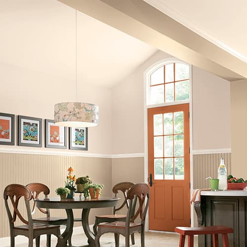 Bright dining area with wooden round table and chairs, floral pendant light, artwork on the wall, and an orange door with window panes.