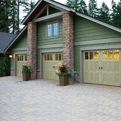 Green house exterior with three beige garage doors framed by stone columns and potted plants on a paved driveway.