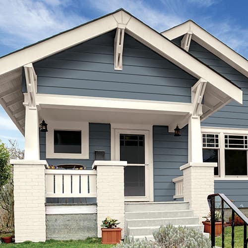 Front porch of a blue house with white trim, white brick pillars, and potted plants on either side of concrete steps.