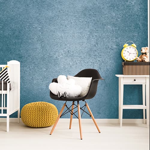 Modern nursery room with a black chair holding white pillows, a yellow knitted pouf, a white crib, and a white side table with a yellow alarm clock and teddy bears against a blue textured wall.
