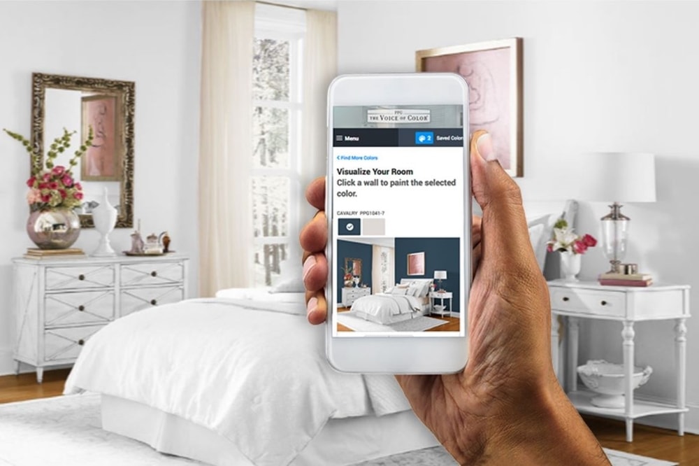 Hand holding smartphone displaying a room color visualizer app against a bedroom background with white bedding and light-colored furniture.