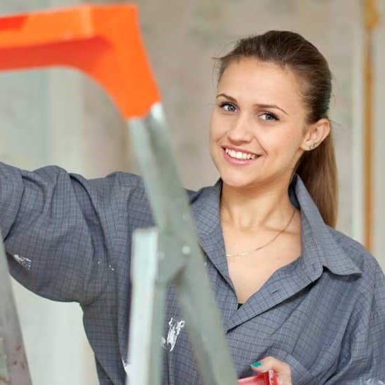 Smiling woman in a grey checkered shirt standing behind a ladder, holding a paint tool.