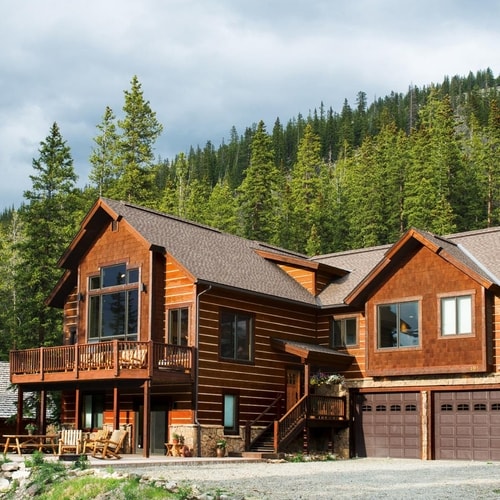 Large wooden cabin with deck and multiple windows surrounded by pine trees on a cloudy day.