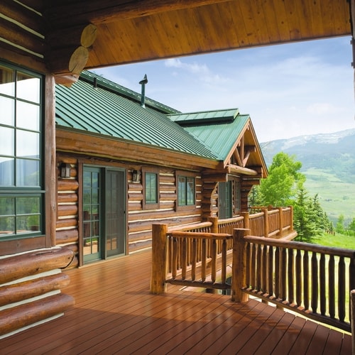 Spacious wooden deck of a log cabin with green metal roof overlooking a scenic mountain landscape with trees and grassy hills.