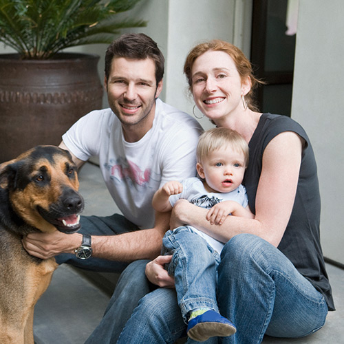 Smiling man and woman sitting with a toddler and a large dog in a casual home setting.