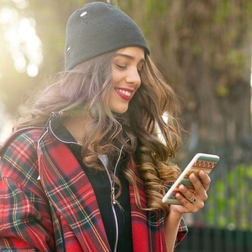 Young woman in a black beanie and red plaid jacket smiling while looking at her phone outdoors.