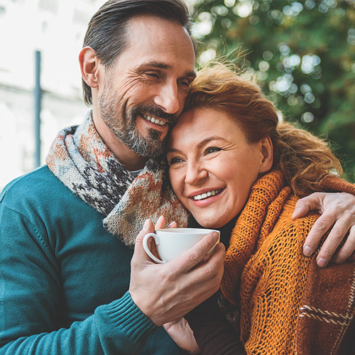 Smiling couple embracing outdoors, the man holding a white cup and both wearing warm scarves.