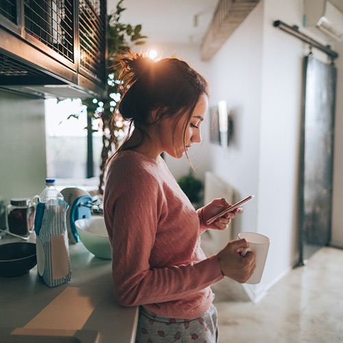 Woman in pink sweater holding a white mug and looking at her phone in a bright kitchen.