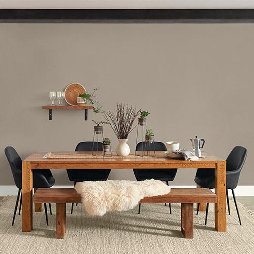 Modern dining room with wooden table, bench with white fur throw, four black chairs, and minimalist decor on taupe wall.