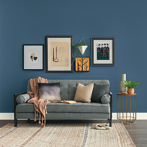 Modern gray sofa with assorted pillows and a beige throw blanket against a dark blue wall with framed abstract and botanical artwork, next to a round side table holding plants and vases.