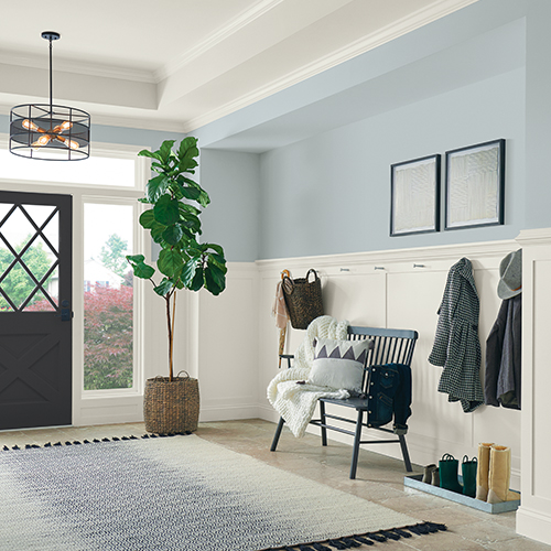 Bright entryway with a black door, a large potted plant, framed art on wall, a bench with throw blankets and a pillow, and coats hanging on hooks.