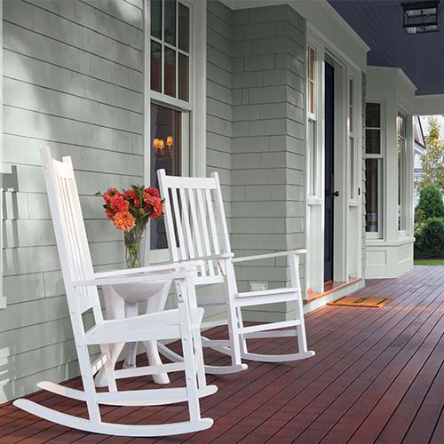 Two white rocking chairs and a small white table with a vase of red and orange flowers on a wooden porch outside a gray house.