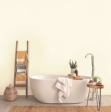 Modern white freestanding bathtub with a towel draped over the edge, a wooden ladder with hanging towels, and potted plants on a small round side table and bathtub tray in a minimal bathroom.