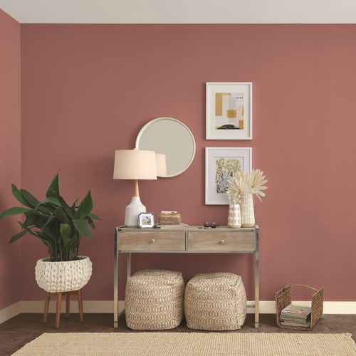 Living room corner with a dusty rose wall, a small wooden console table with a lamp, two poufs underneath, a plant in a woven basket, a round mirror, two framed artworks, and a magazine holder.