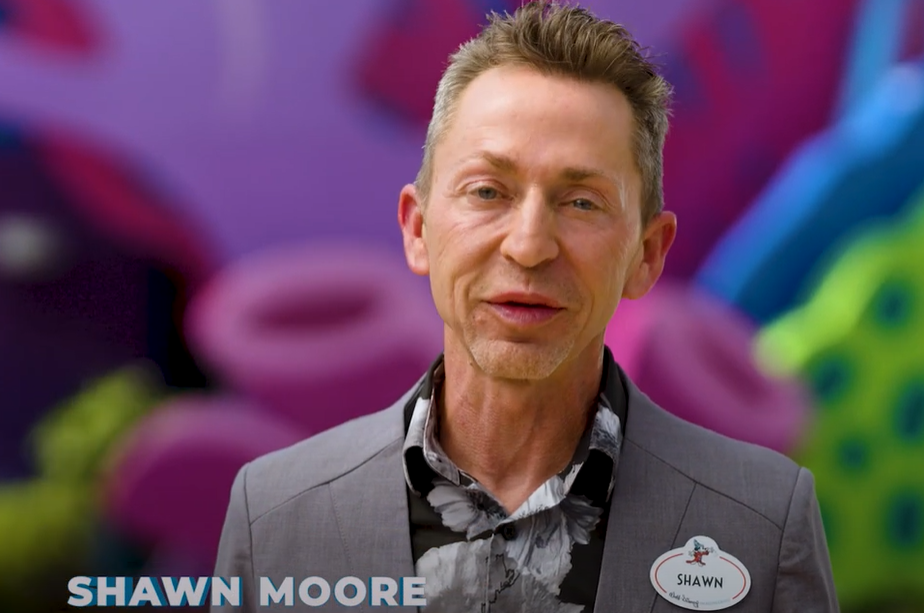Man wearing a suit jacket and patterned shirt with a name tag that reads 'Shawn' against a colorful, blurred background.