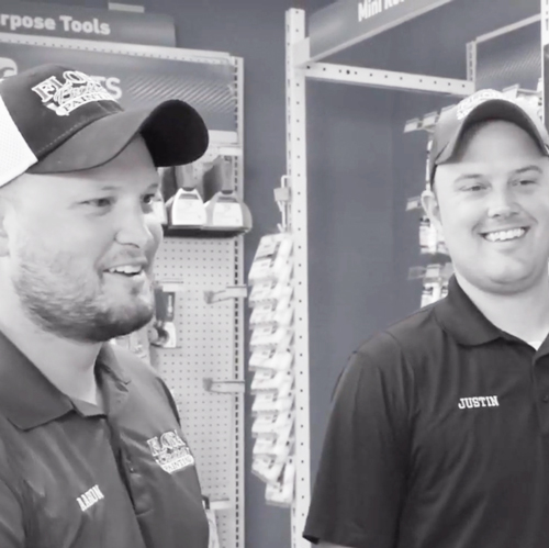 Two men wearing caps and black polo shirts with name tags smiling inside a hardware store aisle.