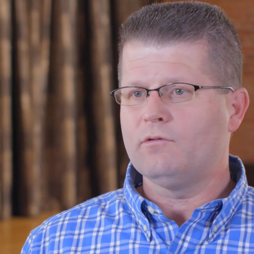 Man with short brown hair and glasses wearing a blue checkered shirt, speaking indoors with blurred brown curtains in background.