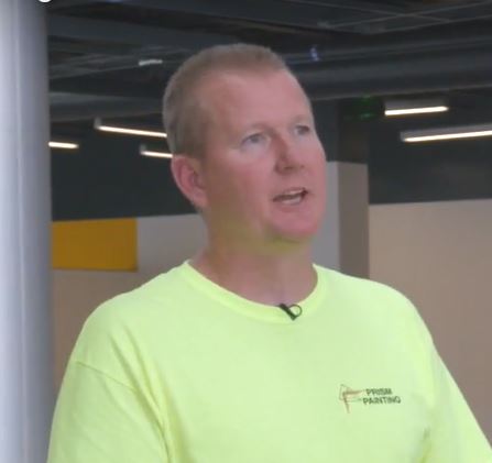 Man wearing a bright yellow Prism Painting t-shirt speaking indoors with ceiling lights in the background.