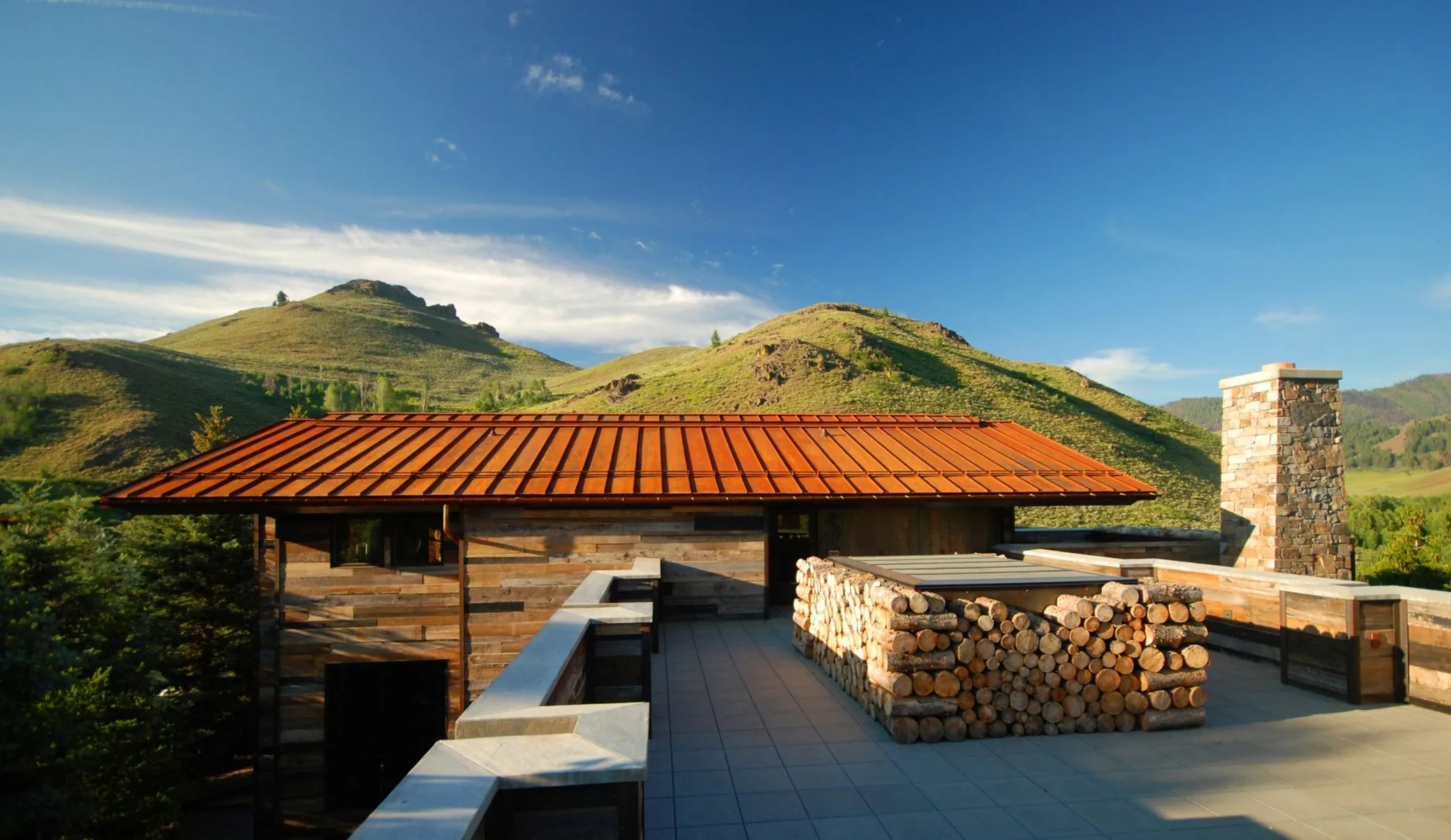 Contemporary mountain home with a rust-colored roof, stone chimney, and stacked firewood terrace, designed by Susan Desko in Lake Creek, Idaho.