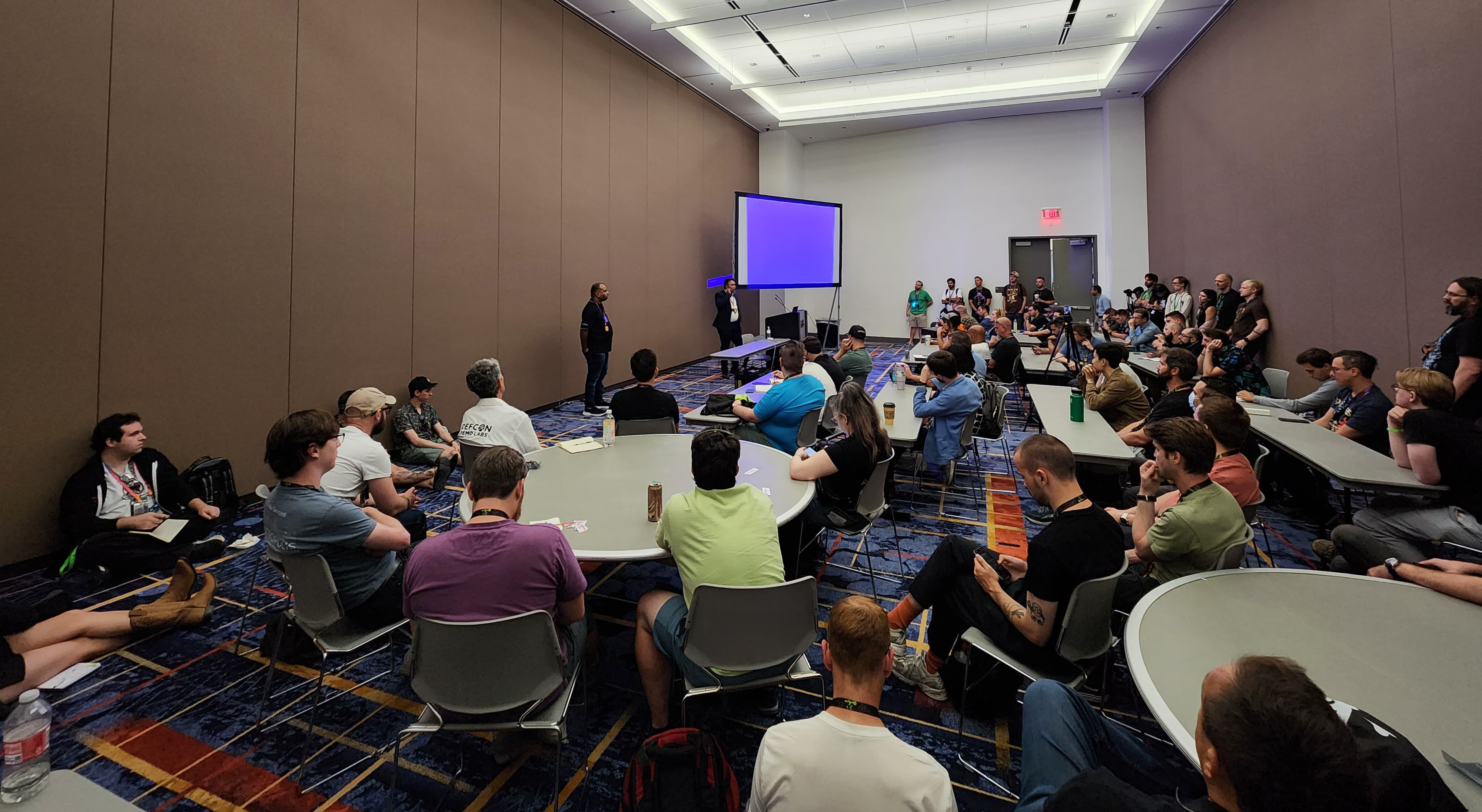 A room full of attendees participating in a Telecom Village workshop, with speakers presenting telecom security concepts.