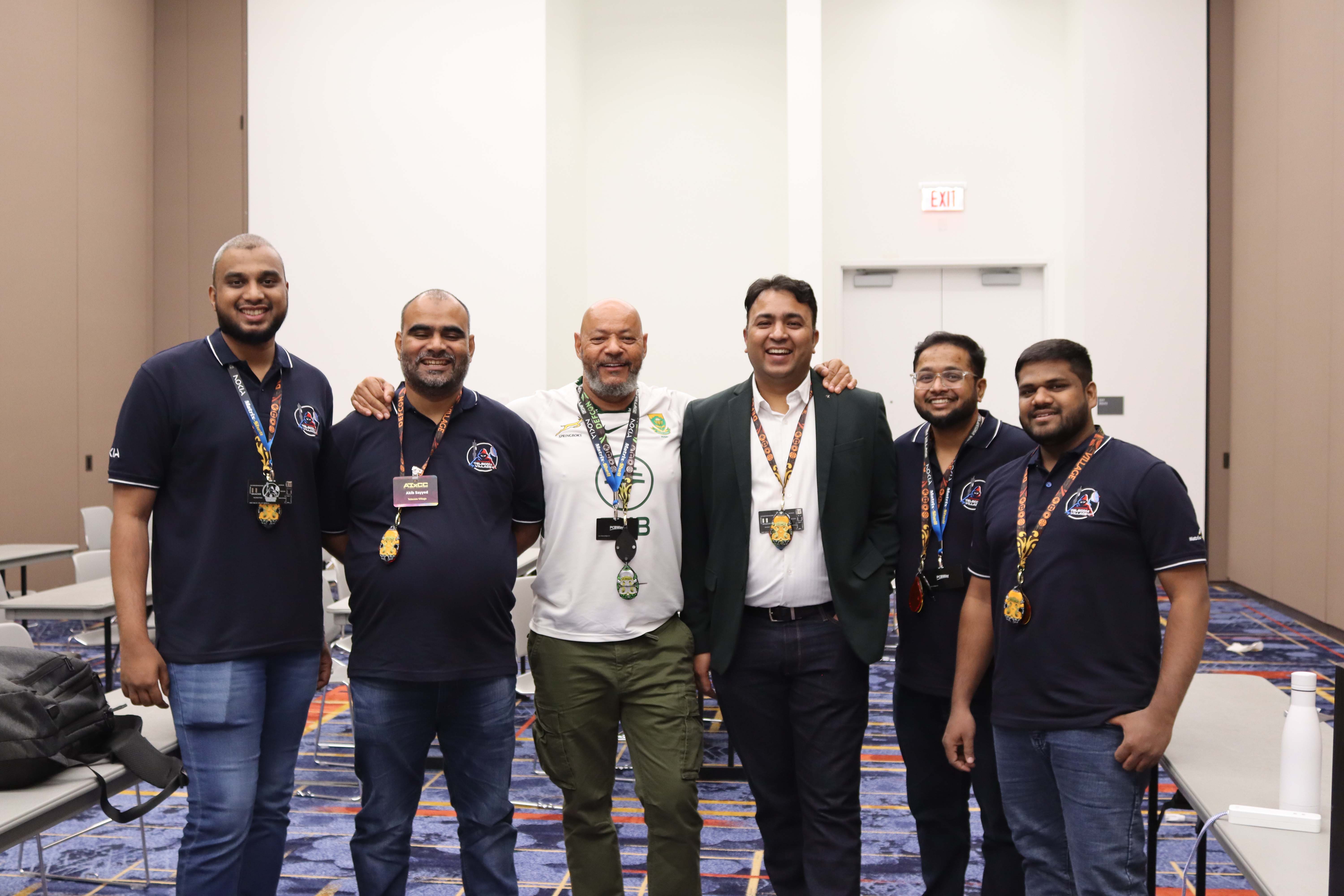 Group of six men standing together indoors, smiling and wearing event badges and lanyards.
