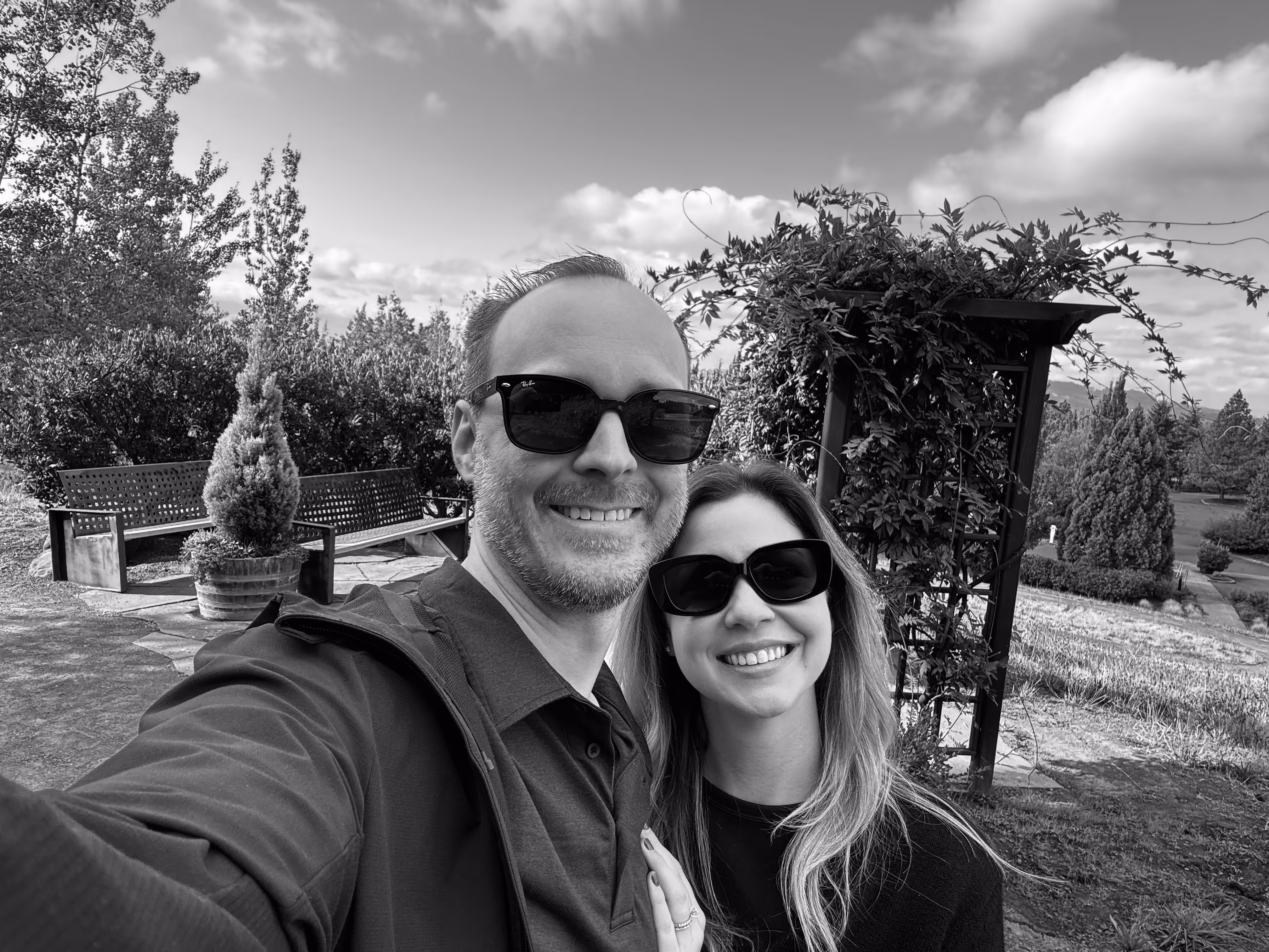 Smiling couple wearing sunglasses outdoors in a garden with benches and greenery behind them.
