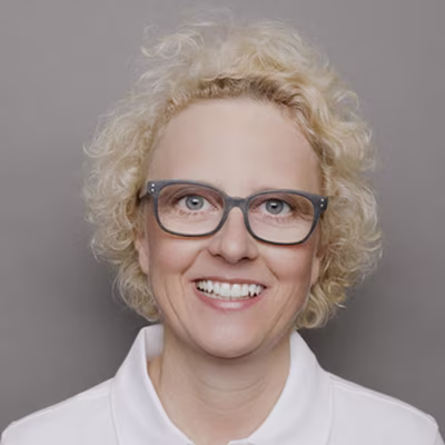 Lächelnde Frau mit lockigem blonden Haar, Brille und weißem Hemd vor grauem Hintergrund.