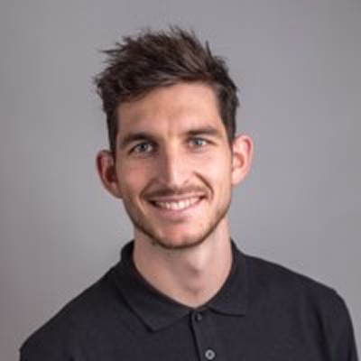 Junger Mann mit kurzen braunen Haaren und Bart, lächelnd im schwarzen Poloshirt vor grauem Hintergrund.