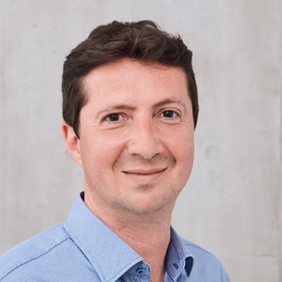 Mann mit kurzen dunklen Haaren im hellblauen Hemd, lächelnd vor hellgrauem Hintergrund.