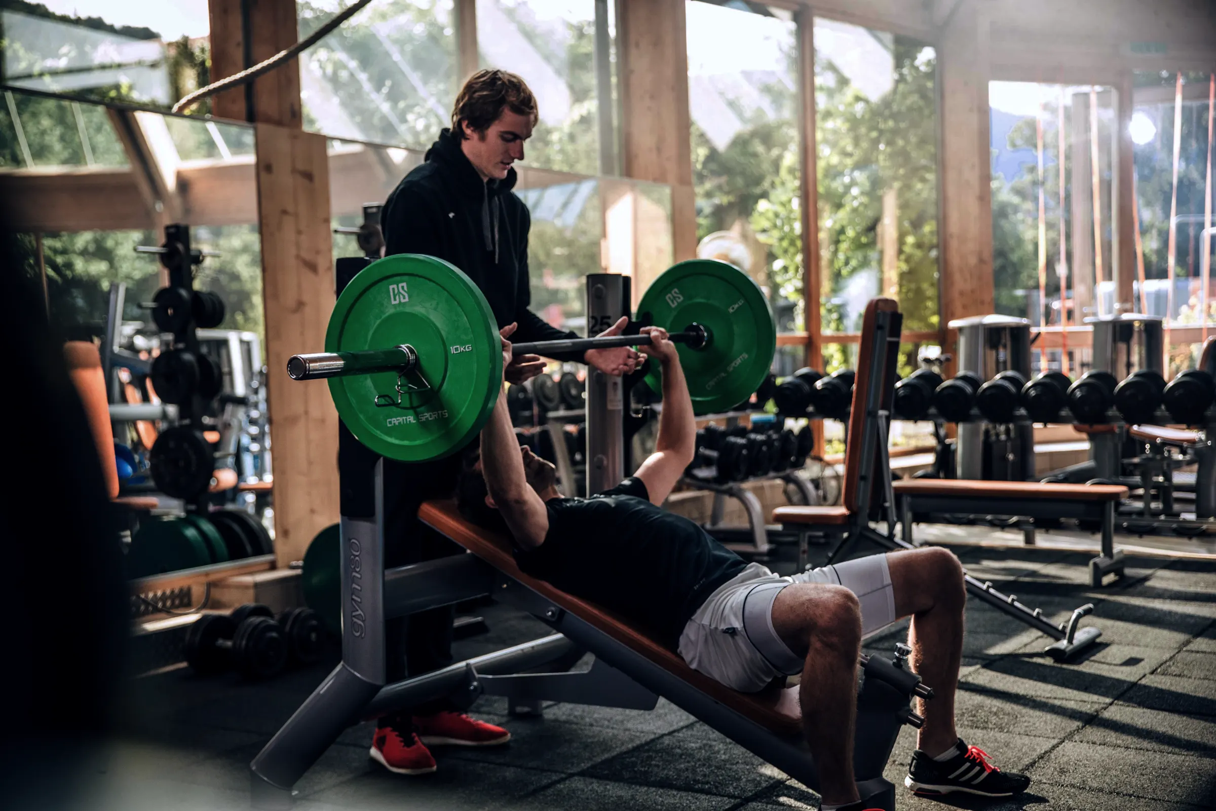 Ein Mann liegt auf einer schrägen Trainingsbank und hebt eine Langhantel mit grünen 10-kg-Gewichtsscheiben, während ein zweiter Mann ihn im hellen Fitnessstudio aufmerksam beobachtet und unterstützt.