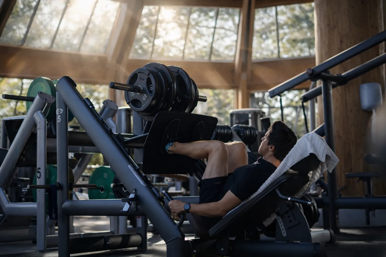 Mann trainiert im Fitnessstudio an einer Beinpresse mit mehreren Gewichten, umgeben von Fitnessgeräten und großen Fenstern mit Außenansicht.