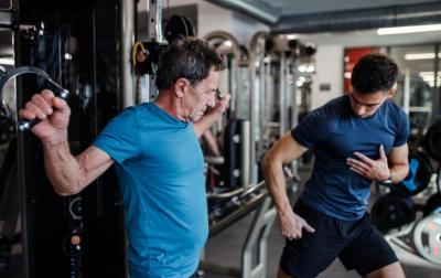 Älterer Mann trainiert im Fitnessstudio am Kabelzug, ein jüngerer Trainer steht daneben und schaut zu