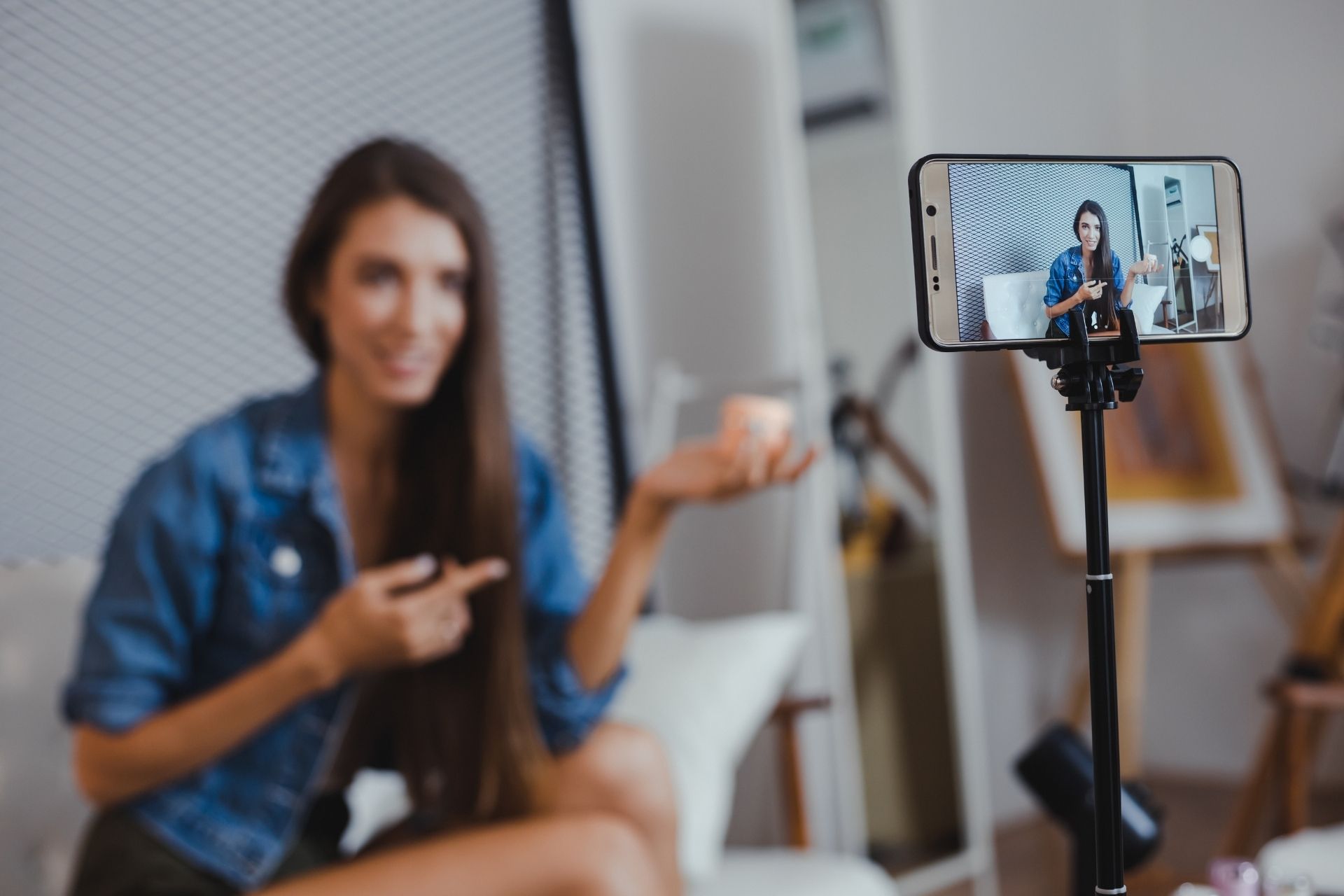 A woman in a denim jacket recording a video on a smartphone mounted on a tripod, pointing at a small container in her hand.