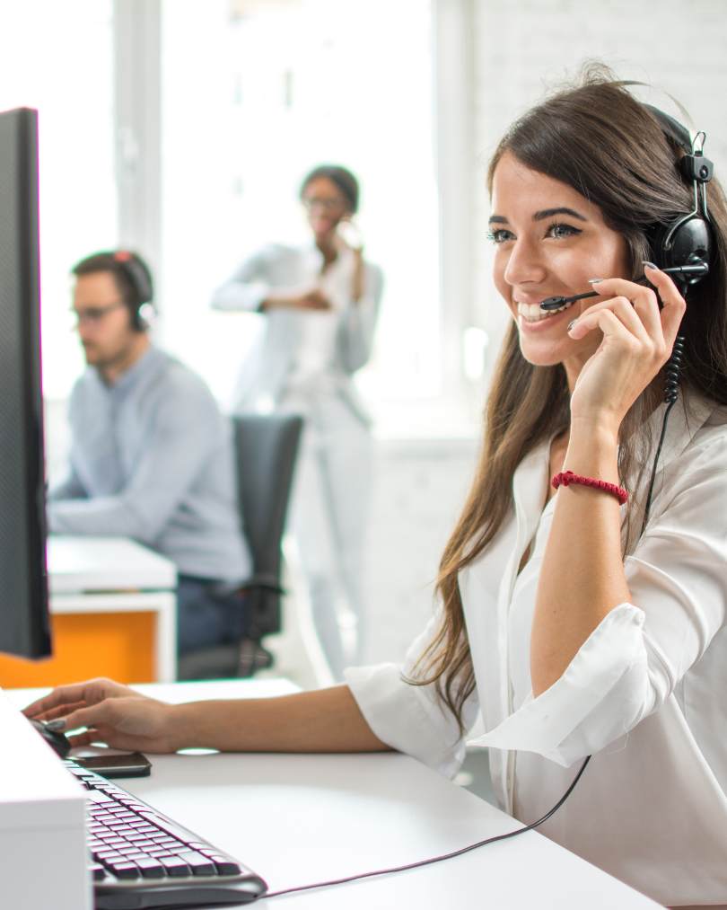 Smiling woman wearing a headset working at a computer in a bright office with colleagues in the background.