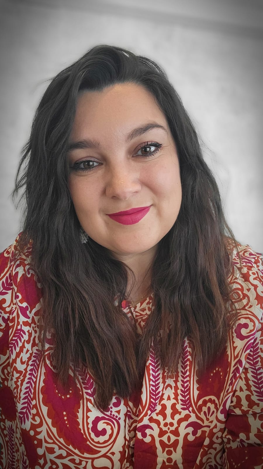 Smiling woman with long dark hair, wearing a colorful red and orange patterned blouse and bright pink lipstick.