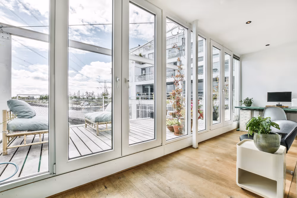 Helles Büro mit Holzboden, großen Glasfenstern zur Terrasse, Pflanzen und Schreibtisch mit Computer.