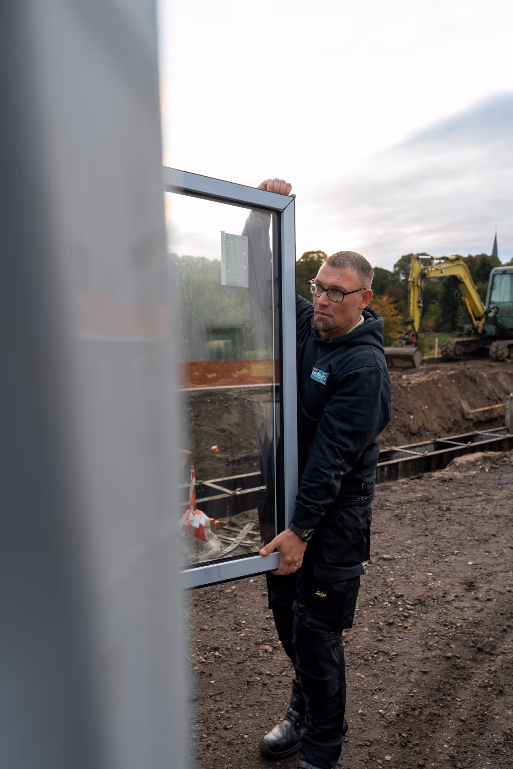 Ein Mann in Arbeitskleidung trägt einen großen Fensterrrahmen auf einer Baustelle mit Baumaschinen im Hintergrund.