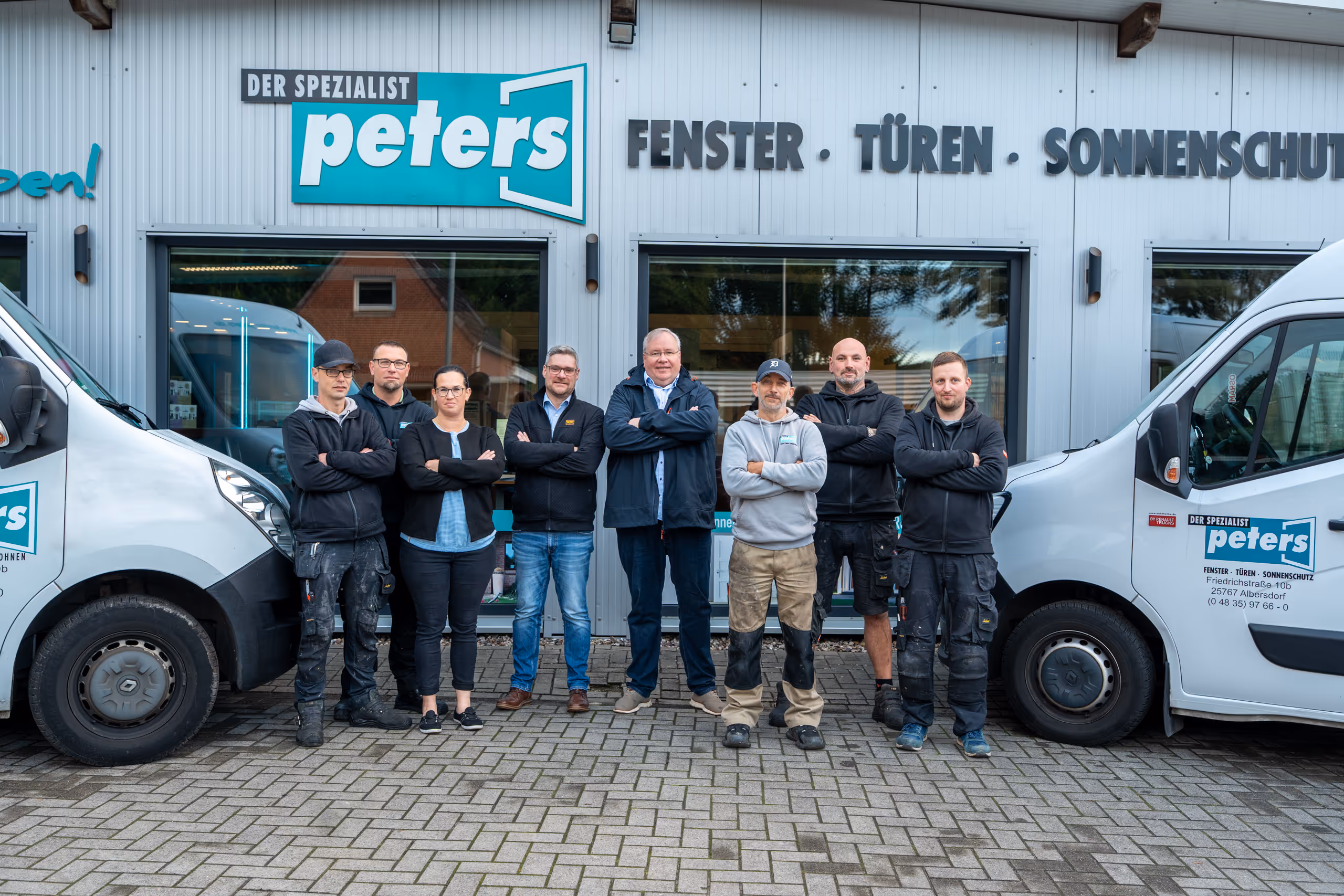 Team von acht Personen steht mit verschränkten Armen vor einem Geschäft mit dem Schild 'Der Spezialist Peters Fenster Türen Sonnenschutz' zwischen zwei weißen Firmenfahrzeugen.
