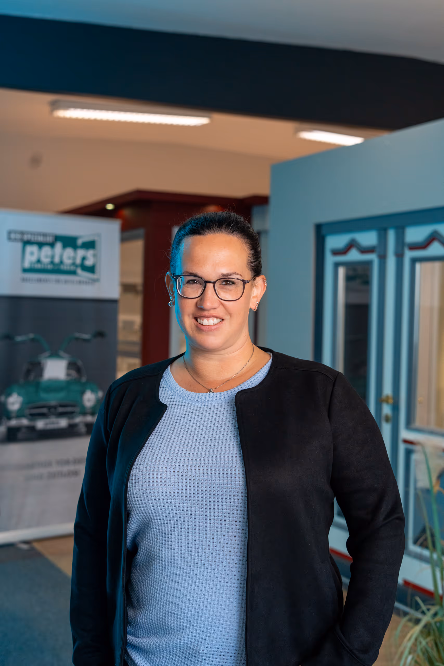 Frau mit Brille, schwarzer Jacke und blauem Pullover lächelt in einem Büro.
