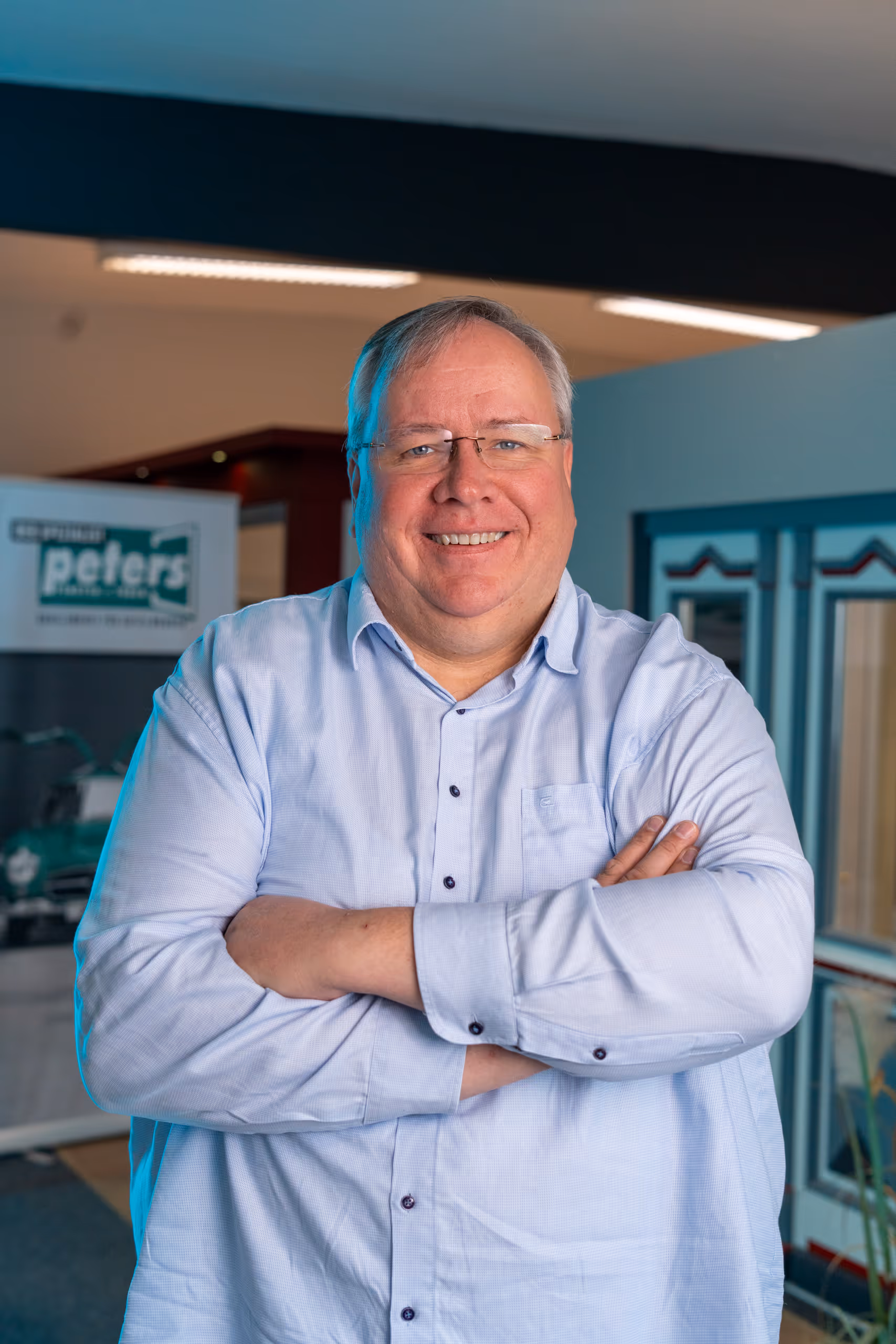 Lächelnder Mann mit Brille und hellblauem Hemd, die Arme vor der Brust verschränkt, vor unscharfem Hintergrund mit einem Schild.