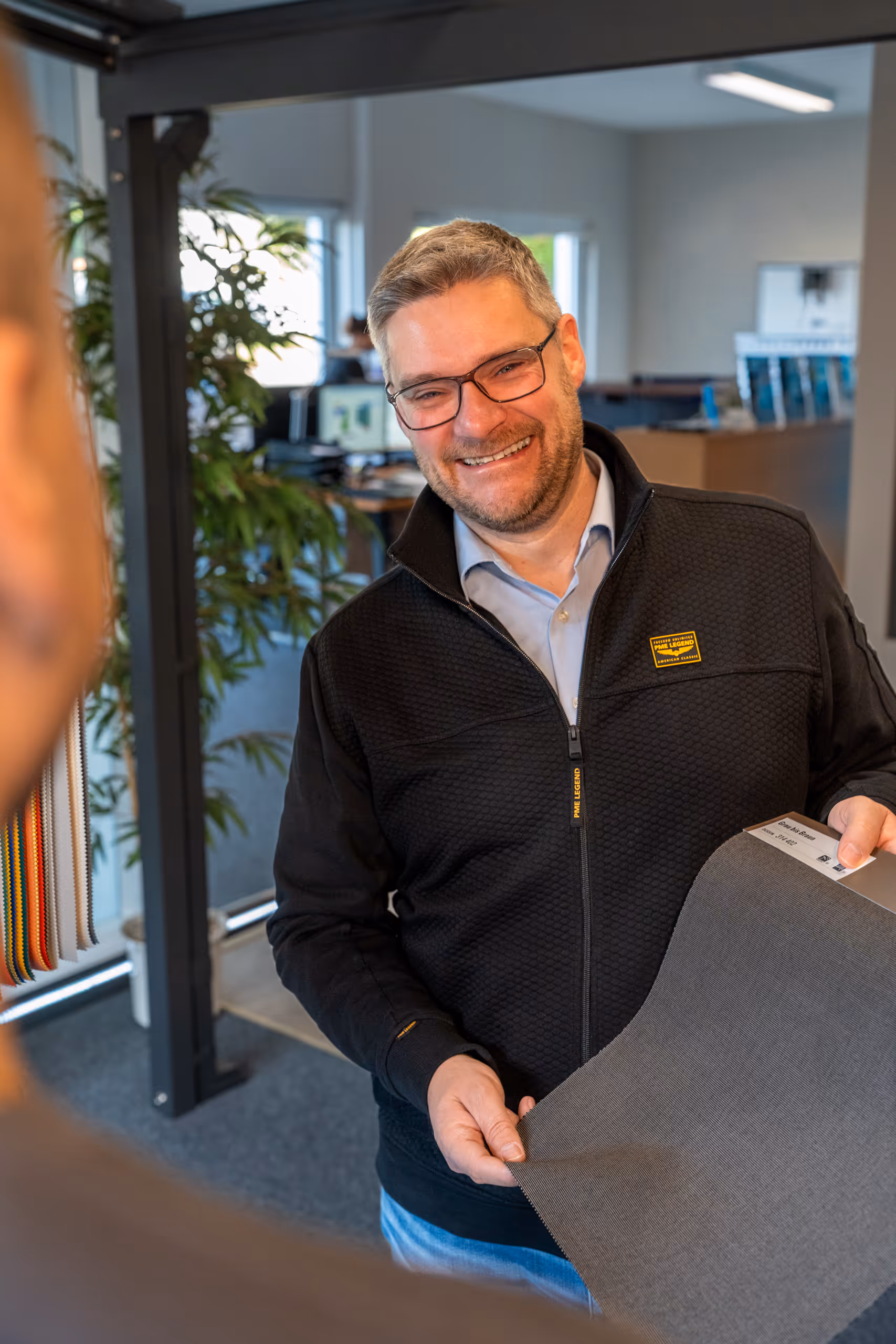 Ein lächelnder Mann mit Brille hält ein Stoffmuster in einem Büro.