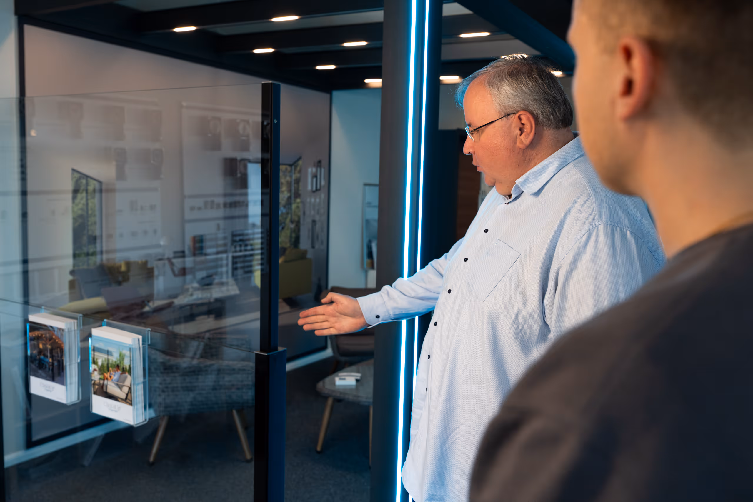 Älterer Mann zeigt auf eine Glaswand in einem modernen Büro, während eine andere Person im Vordergrund zuhört.
